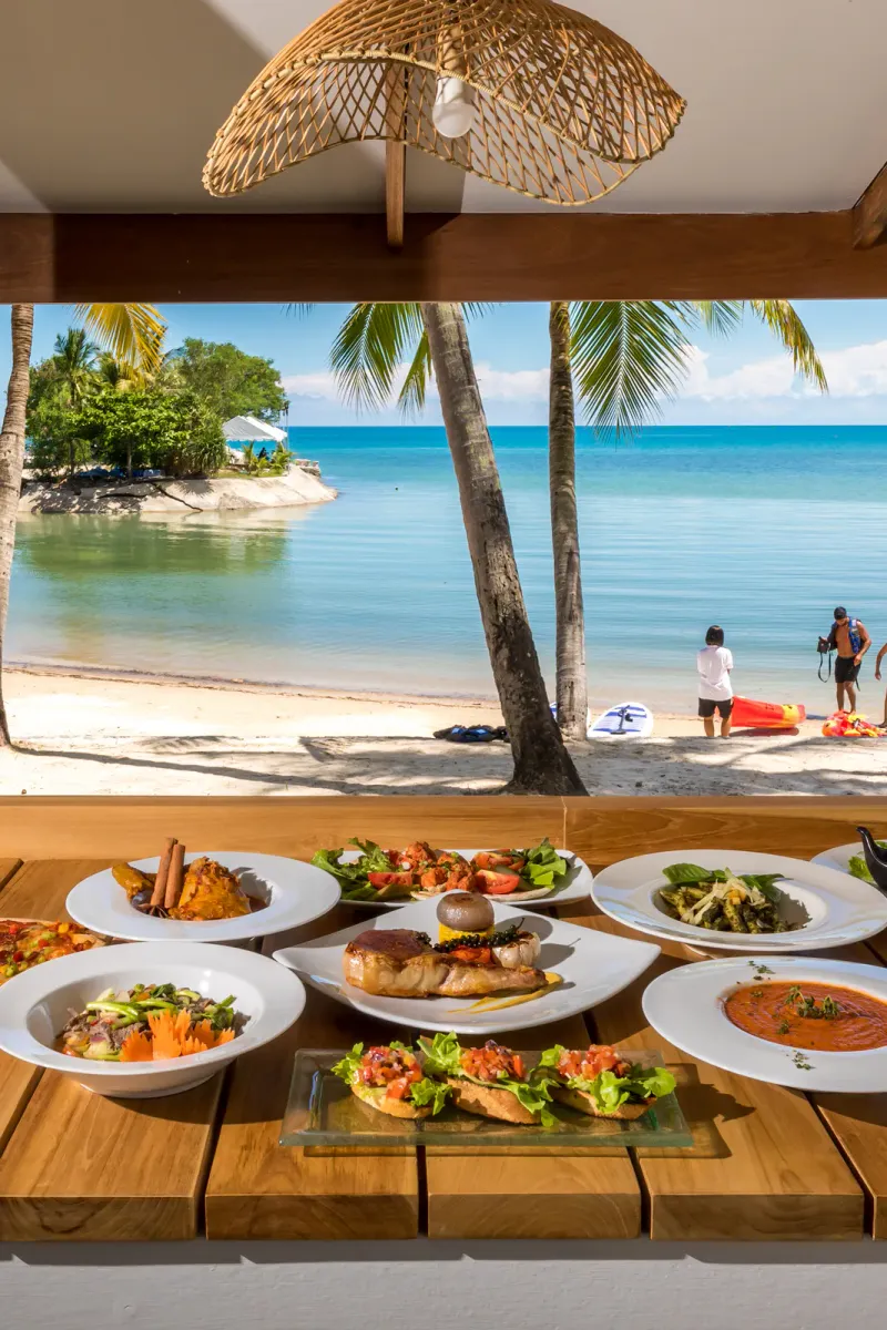 Spiaggia di sabbia fiancheggiata da palme con kayak e un tavolo di piatti thailandesi dalla pagina cibo e bevande dell'Outrigger Koh Samui