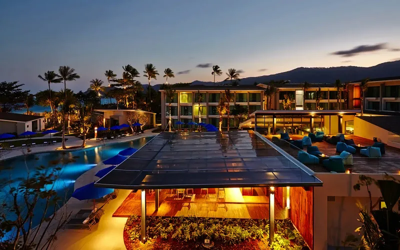 Veduta al crepuscolo della piscina dell'OZO Chaweng Samui, del padiglione del bar illuminato e dei blocchi di camere sotto le palme sulla spiaggia di Chaweng