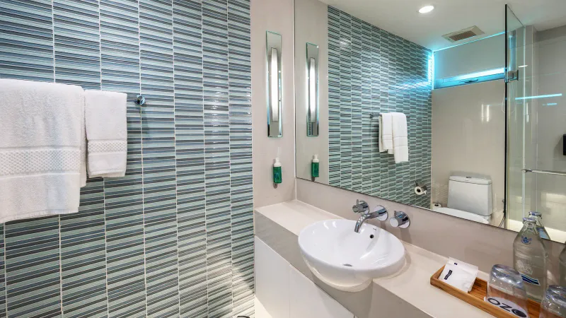 Bagno dell'OZO Chaweng Samui con una parete di piastrelle di vetro grigie, lavabo bianco, asciugamani e una doccia di vetro