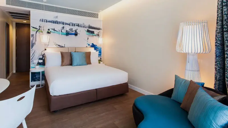 Letto matrimoniale in una camera dell'OZO Chaweng Samui con un murale di barca da pesca, cuscini turchesi e una lampada bianca
