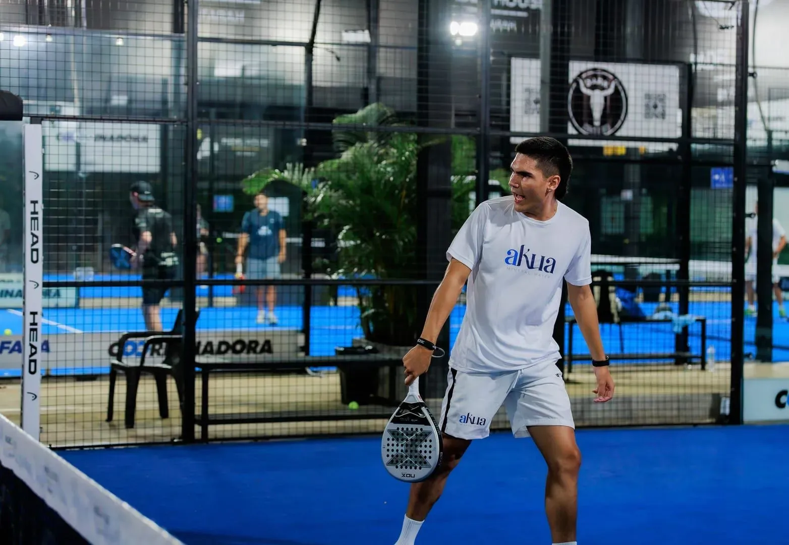 Padel player mid-swing on an indoor court, the fast rally style that fills a club's weekly social night