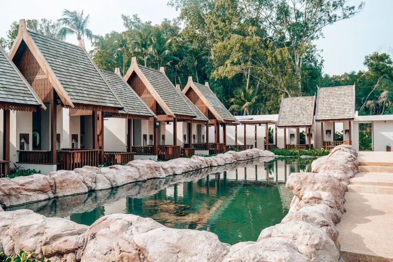Rangée de pavillons en bois de style thaï à toits à deux pans au bord d'un bassin aux rives de pierre dans les jardins du resort