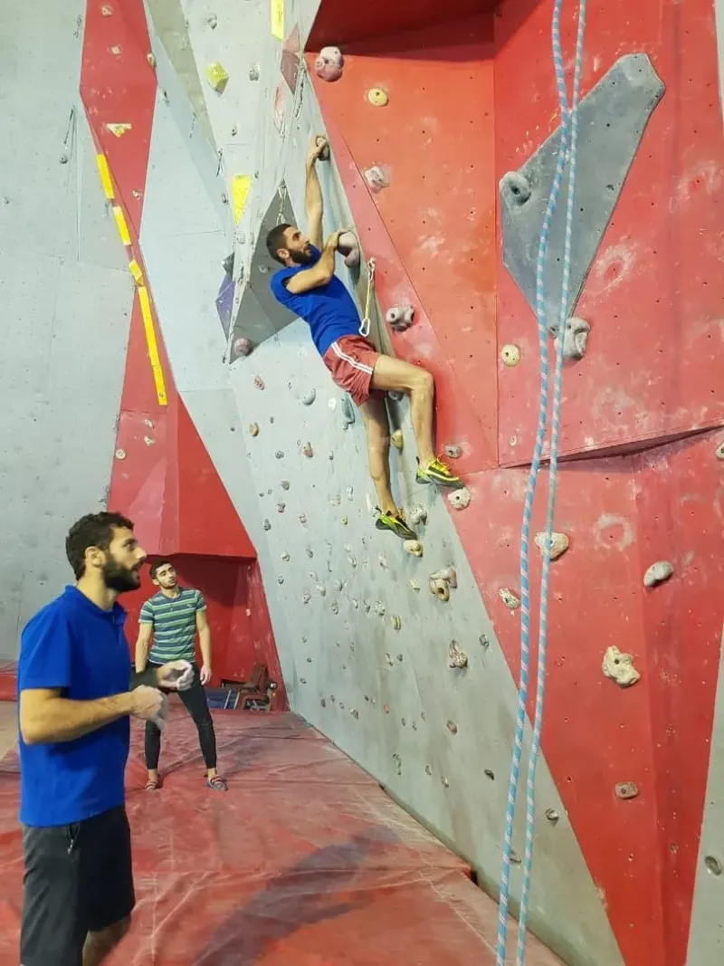 Climber on a roped indoor wall with a belayer watching from the mat below, the belay-partner setup Samui lacks
