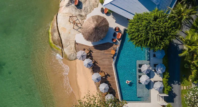 Veduta aerea di una terrazza con piscina blu e ombrelloni accanto alle rocce e a una baia di sabbia al Rocky's Boutique Resort