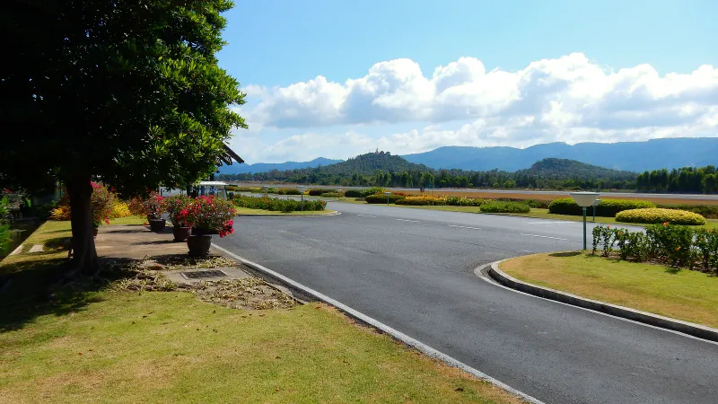 Strada con giardini accanto alla pista dell'aeroporto di Samui, con colline verdi e un tempio in cima sullo sfondo