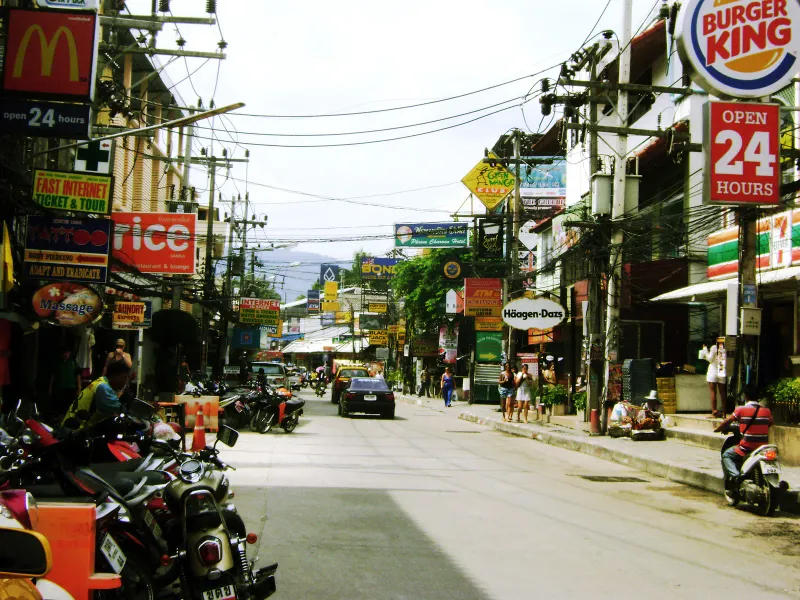 Rue animée de Chaweng avec des scooters garés, des enseignes de boutiques et un taxi au loin à Koh Samui