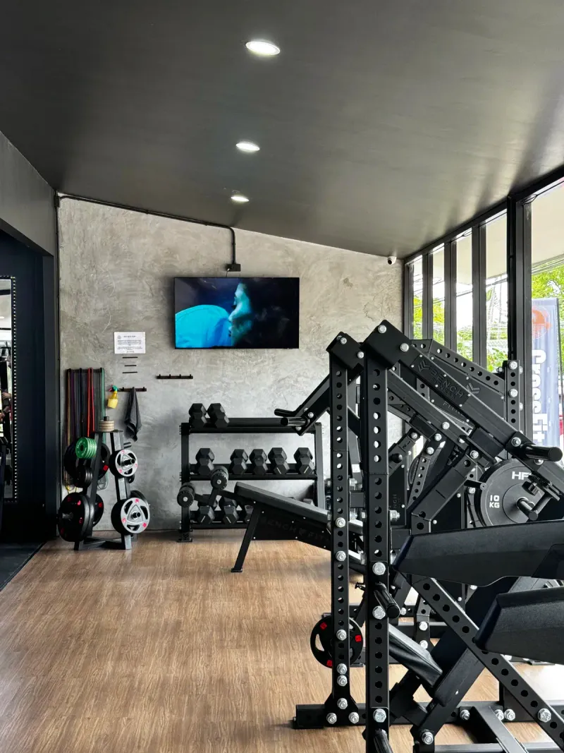 A gym room with racks, dumbbells and a wall-mounted screen beside tall windows