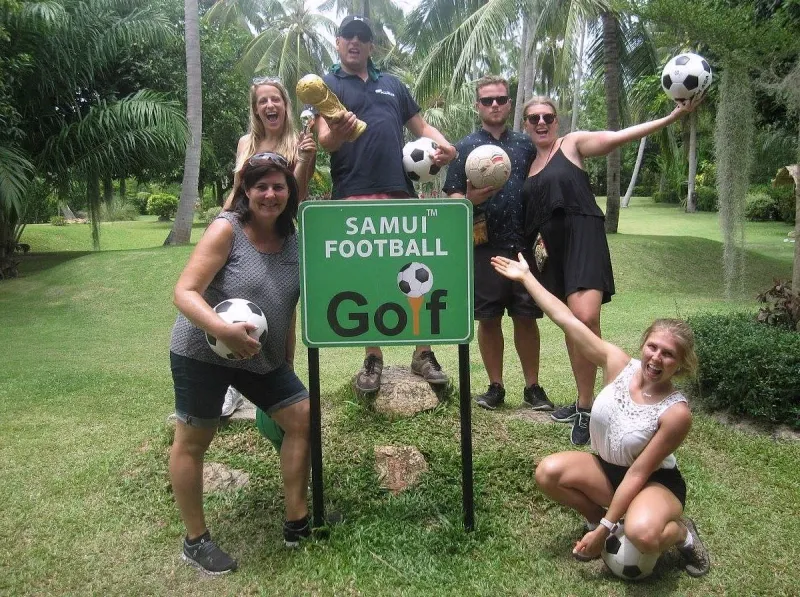 Visitors posing beside the Samui Football Golf course sign among the palms
