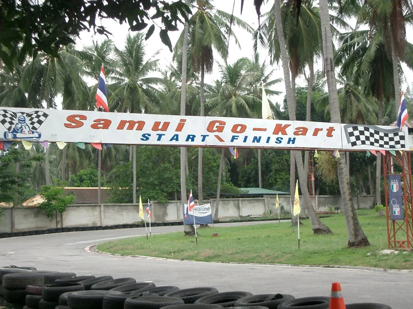 Weathered start/finish banner reading Samui Go-Kart over an empty asphalt track lined with tyre barriers, Bophut