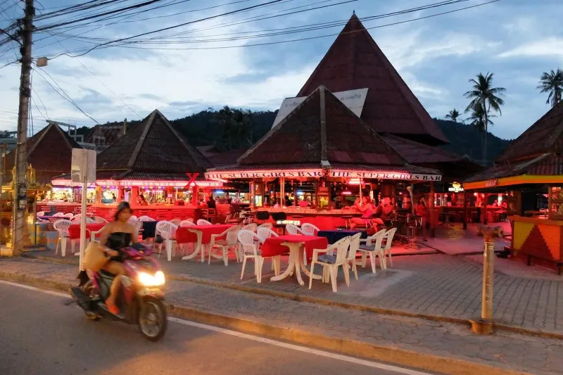 Beachfront bars and restaurants lit in red at dusk beside a coastal road on Koh Samui
