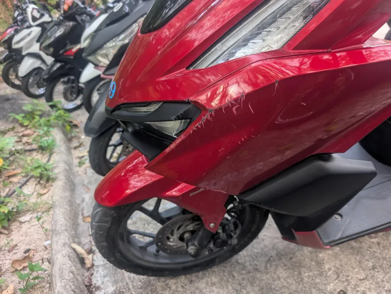 Carena anteriore rossa di uno scooter graffiata, con la cornice del faro staccata, accanto a scooter parcheggiati