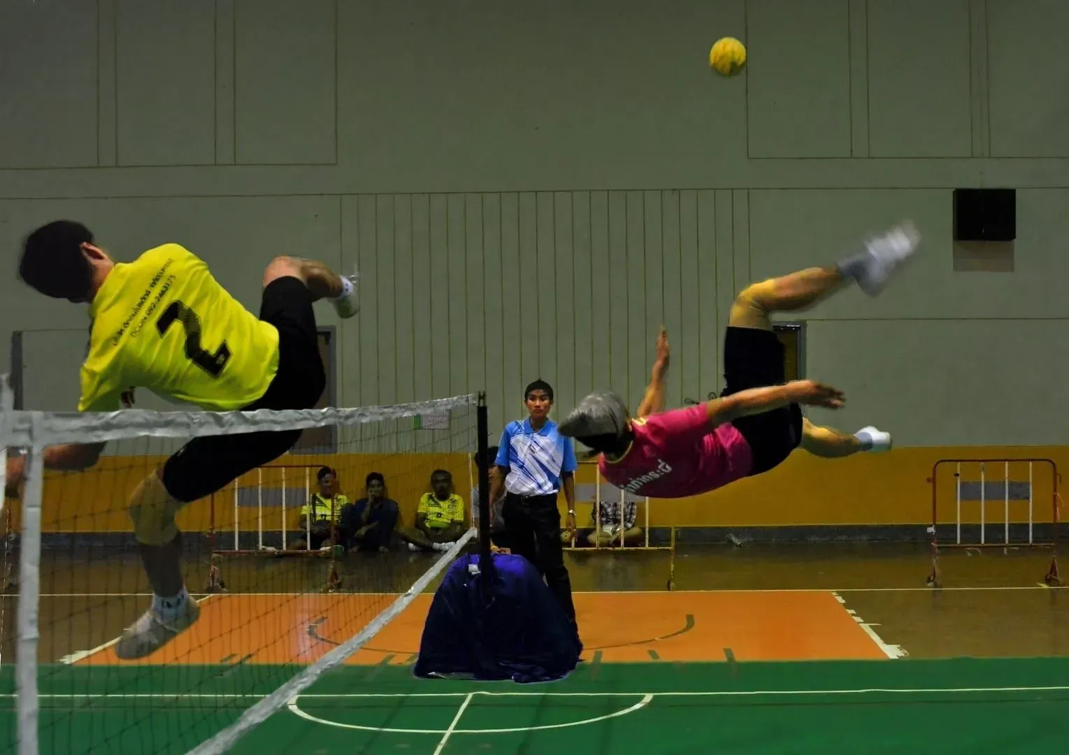A sepak takraw player leaps backward over the net for an overhead kick during a match, teammates and a referee looking on