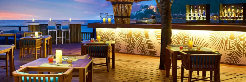 Terrazza del beach bar Blue Monkey al tramonto, con tavoli a lume di candela e un bancone con stampa di palme allo Sheraton Samui Resort