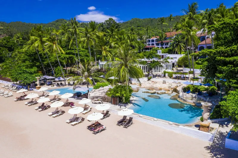 Veduta aerea della piscina fronte spiaggia dello Sheraton Samui Resort, con lettini sulla sabbia e la collina boscosa con gli edifici bianchi del resort
