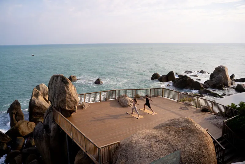 Due persone praticano yoga su una piattaforma di legno costruita sui massi di granito sopra il mare