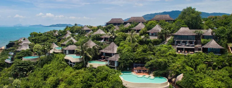 Veduta aerea di ville con tetto di paglia e piscine a sfioro a gradoni su una collina boscosa sopra il mare al Silavadee Pool Spa Resort