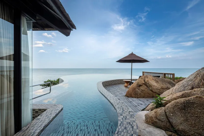 Bordo curvo di una piscina a sfioro accanto a una villa, con massi di granito e mare aperto fino all'orizzonte