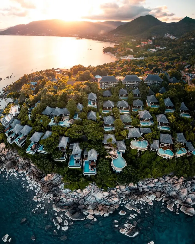 Veduta aerea al tramonto di un resort di ville sulla collina con piscine private su un promontorio roccioso e, più in là, la baia di Lamai