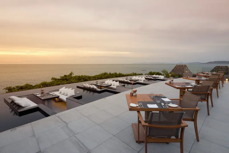 Terrazza all'aperto di un ristorante sul tetto, con sedute incassate e tavoli rivolti verso il mare al tramonto
