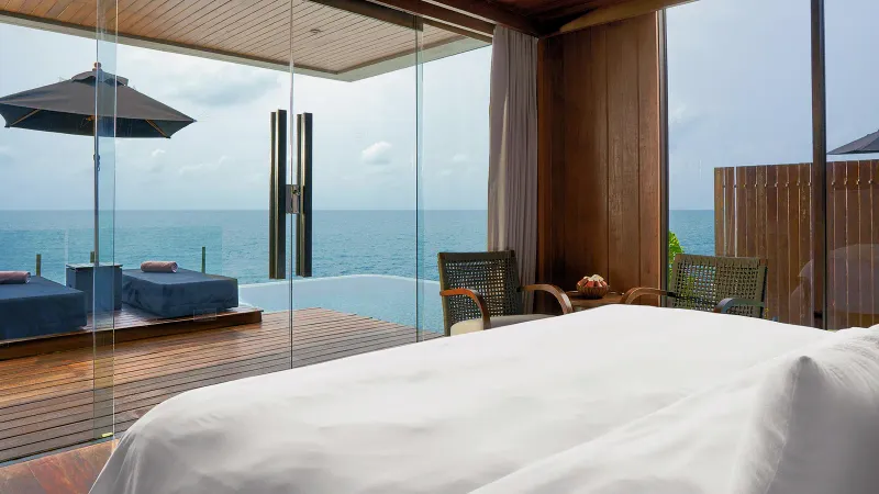 Camera da letto di una villa con lenzuola bianche e una parete di vetro che si apre sul ponte con la piscina privata e sul mare
