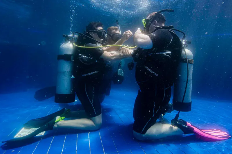 Two scuba students kneeling on the bottom of a training pool with tanks and fins