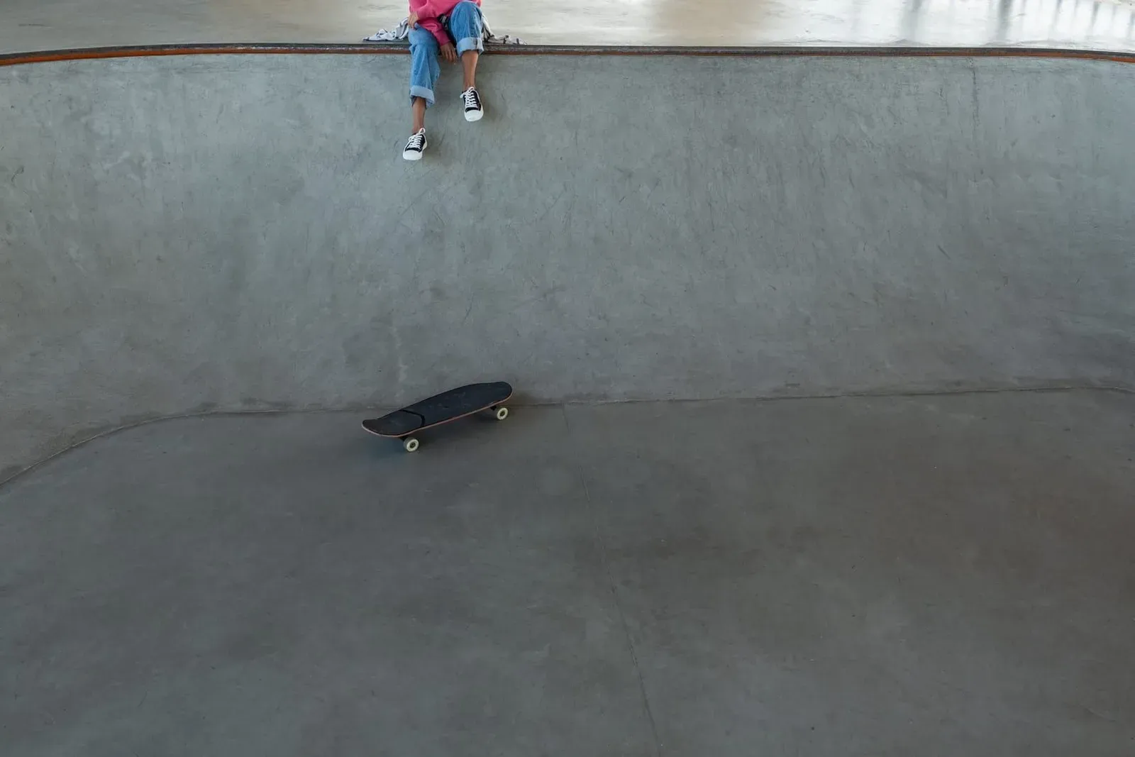 Skateboarder sitting at the edge of a concrete skatepark bowl with the board resting below