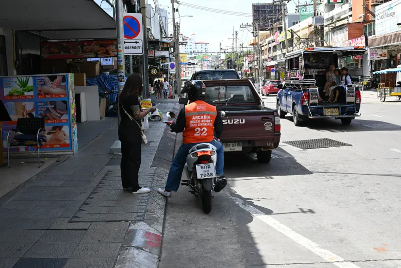 タイの道路脇でソンテウの隣に待機する、オレンジの番号付きベストを着たバイクタクシー運転手