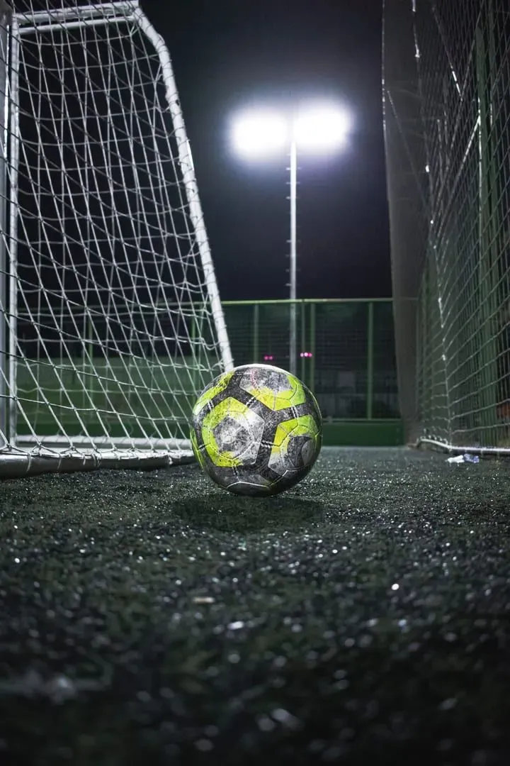 A worn football resting on artificial turf in front of a goal net, floodlights glowing behind it at night