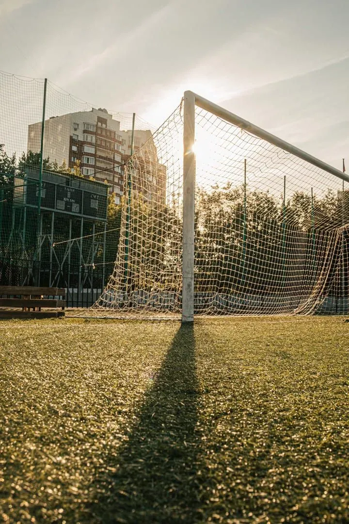 A football goal net backlit by a low evening sun on a turf pitch with apartment buildings behind