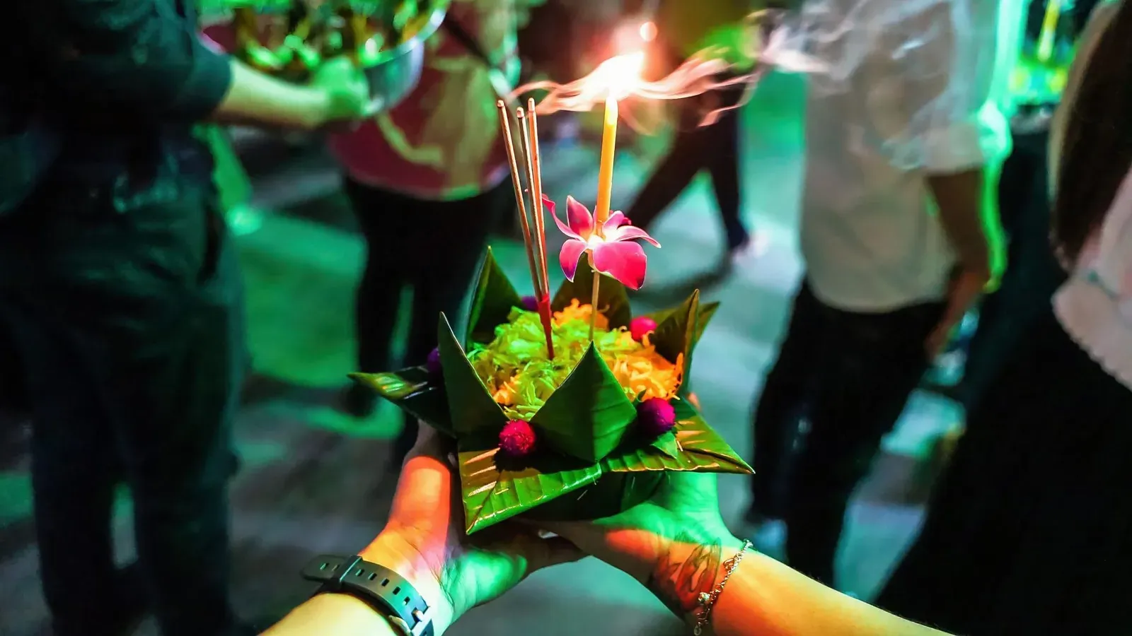 Hands holding a floating krathong offering decorated with a candle and flowers during a Thai festival