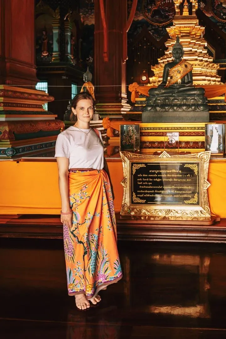 Visitor wearing a colorful sarong inside an ornate Thai temple hall near a Buddha statue