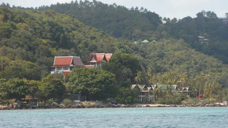 Thai-style pitched roofs among palm trees on a green hillside above a rocky shore, northwest Koh Samui