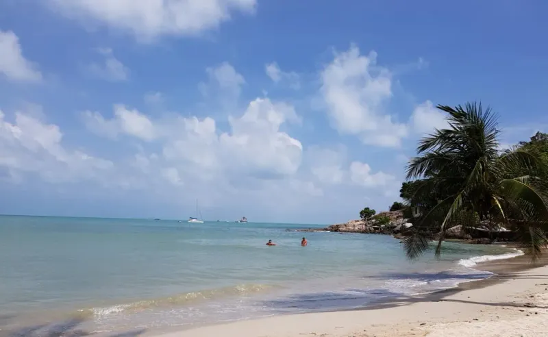 Schwimmer im flachen Wasser neben verankerten Booten vor einem ruhigen Strand auf Koh Samui