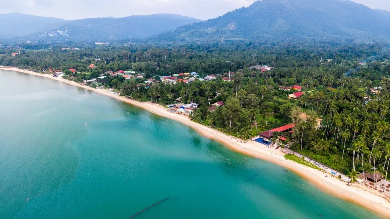 Luftaufnahme einer langen Sandbucht, gesäumt von Palmenhainen und einem Strand-Dorf auf Koh Samui