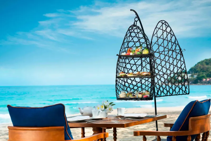 Présentoir à thé de l'après-midi sur une table au bord de la plage à Vana Belle, avec une eau turquoise derrière