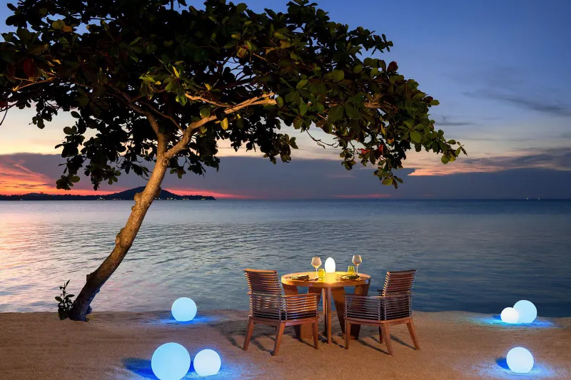 Table pour deux aux chandelles sur le sable à la tombée du soir, avec des sphères lumineuses et la mer derrière