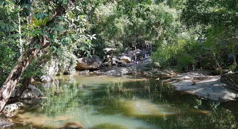 A shaded jungle pool with granite boulders where visitors wade in the shallows