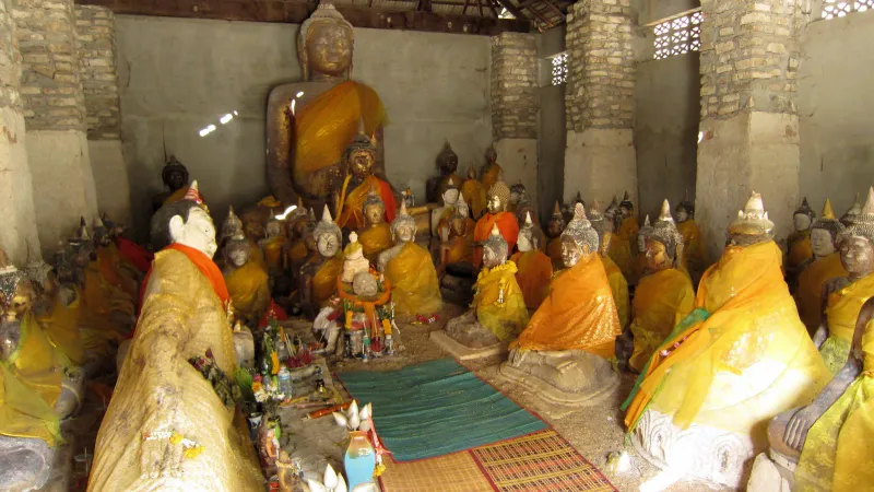 Interior of the weathered brick hall at Wat Samret packed with dozens of seated Buddha statues draped in orange and gold cloth