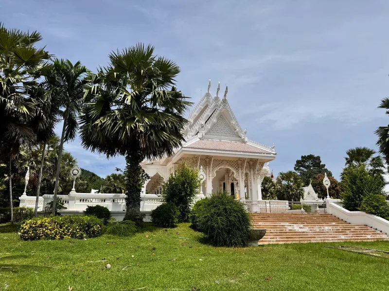 White ordination hall with an ornate carved portico, seen through palm trees on a landscaped lawn