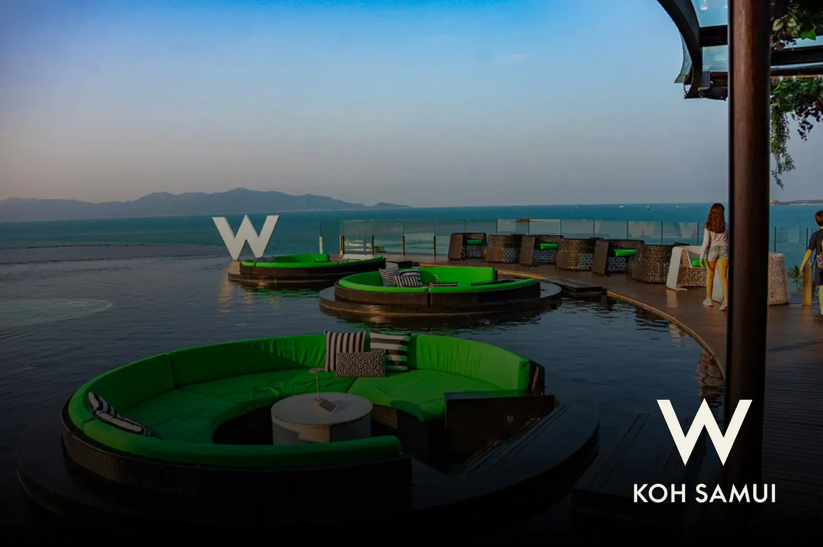WOOBAR's round green daybed platforms on the infinity pool at dusk, with the W Koh Samui sign and Gulf of Thailand view
