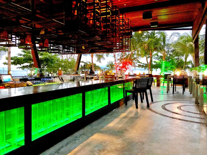 WOOBAR's green-lit outdoor bar counter with palm trees and the sea behind