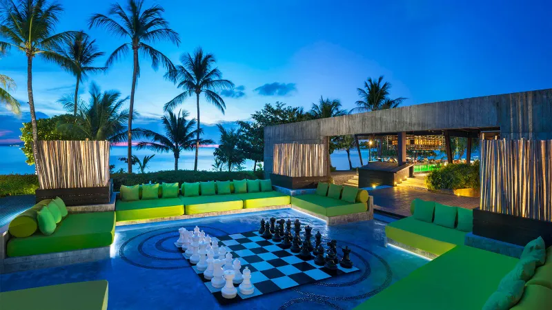 A giant chess set on WOOBAR's lounge deck, ringed by green sofas, at dusk
