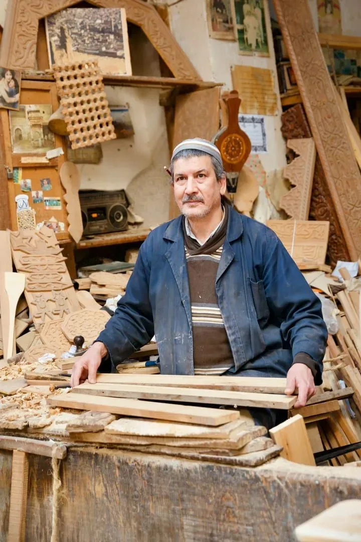 An older craftsman standing in a workshop surrounded by wooden planks and carved pieces