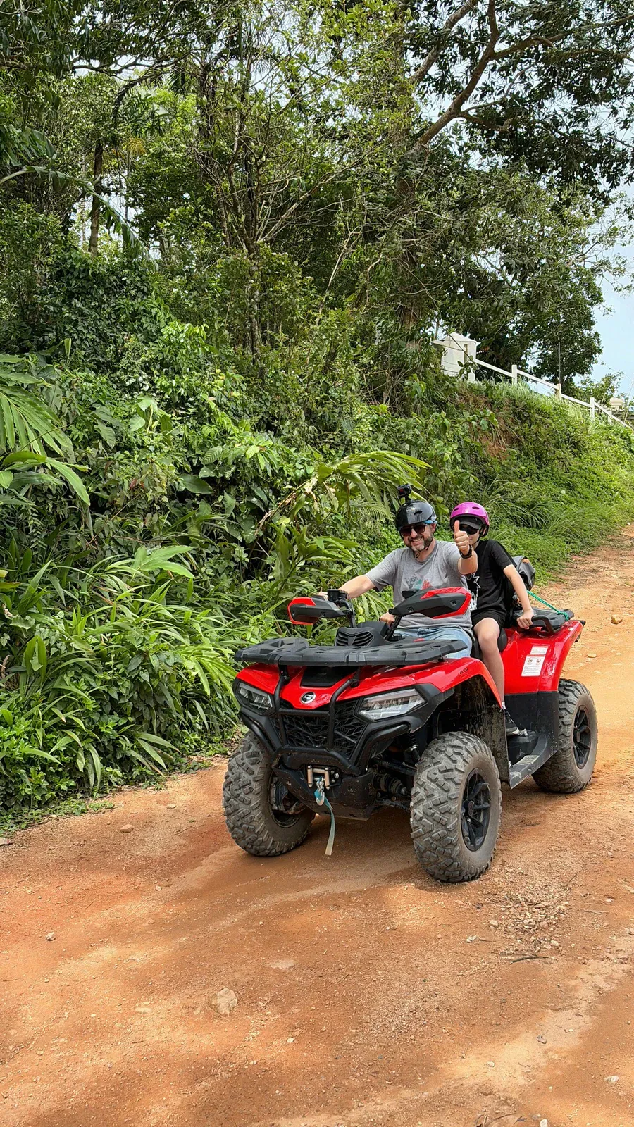 Two riders sharing a quad bike on a red dirt trail with a hillside white fence in the background, X-Quad Samui
