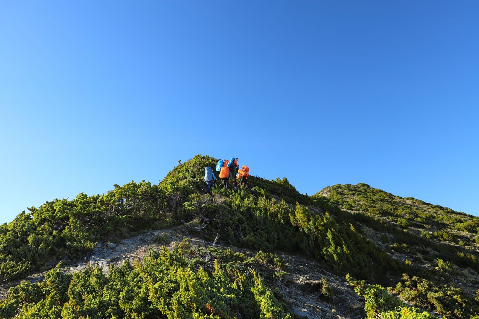 Hikers ascending a lush green mountain ridge trail under blue sky similar to the Maenam Samui traverse